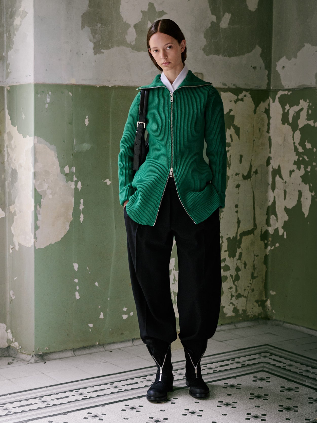 A model stands in a worn interior with peeling green and beige walls, wearing a bright green ribbed zip-front jacket over a white collared shirt, black baggy trousers, and black boots, with a black shoulder bag.