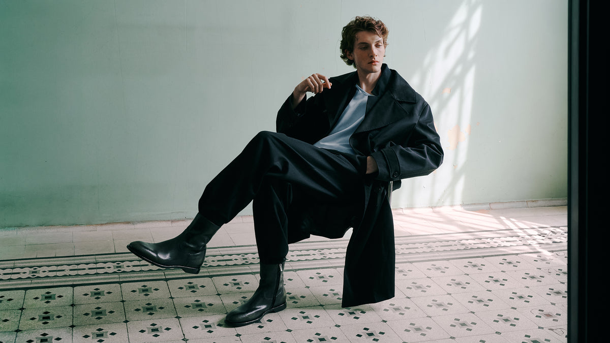 A man wearing black leather boots sits casually on a tiled floor against a pale green wall.