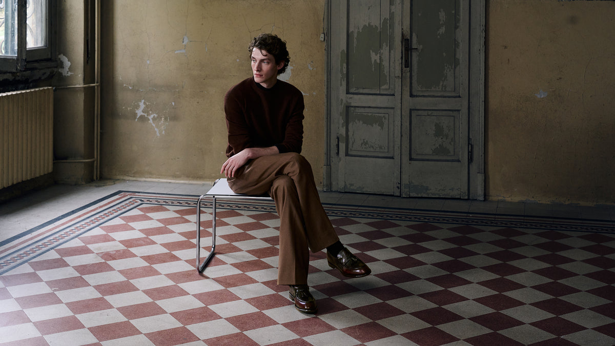 A man sits on a metal chair in a worn interior room with peeling beige walls, a gray door, and a red-and-white checkered floor. He is wearing a dark brown sweater, brown trousers, and a Brown Tassel Loafers.